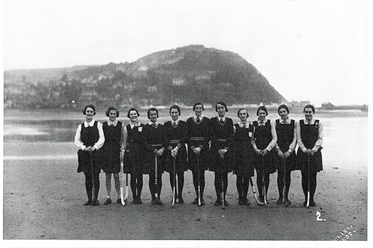 A line of eleven women holding hockey sticks and wearing a mixture of knee-length tunics or sweaters with stockings pose on a beach in front of the sea and a hill atop of spur of land in the background.