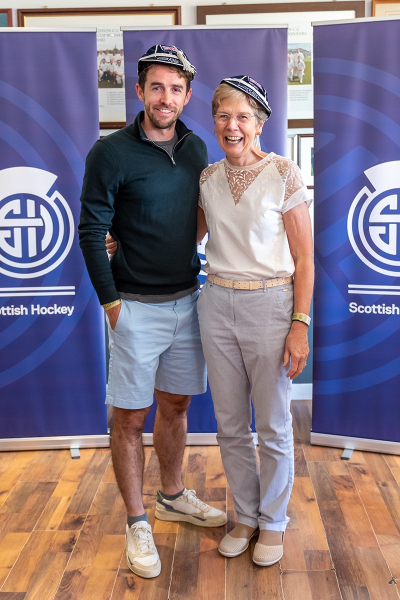 A young man in a long-sleeved, navy blue sweater and light blue shorts stands with an older woman, his mother, who is wearing a white blouse and light blue trousers. They are smiling wearing their Great Britain hockey honours caps which are navy blue with sliver trim.