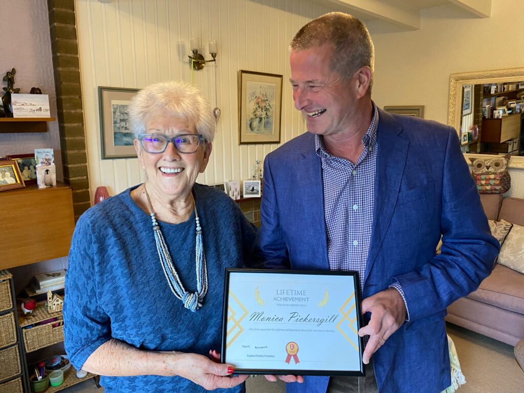 An elderly lady is presented with a framed certificate by a middle-aged man in a living room, both wearing blue