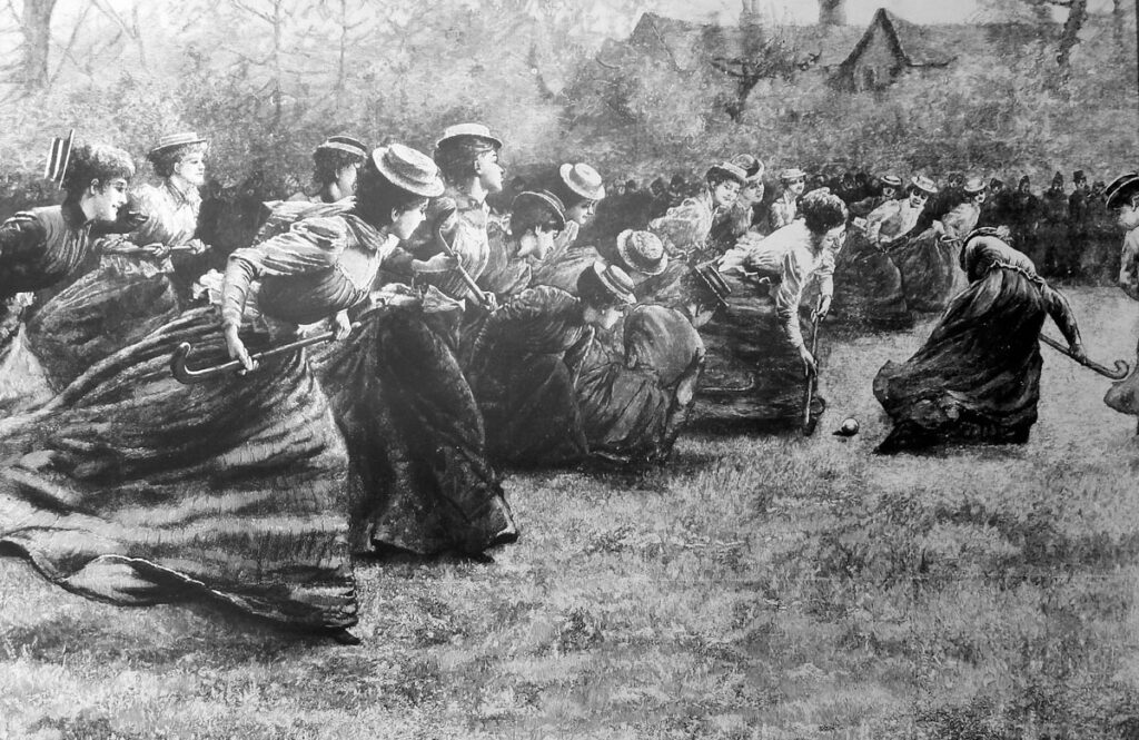 A black and white engraving showing women in ankle-length dresses, blouses and wide-brimmed boater hats playing hockey in a field in front of a row of trees and bushes. The roof of a large house can be seen above the trees in the background.