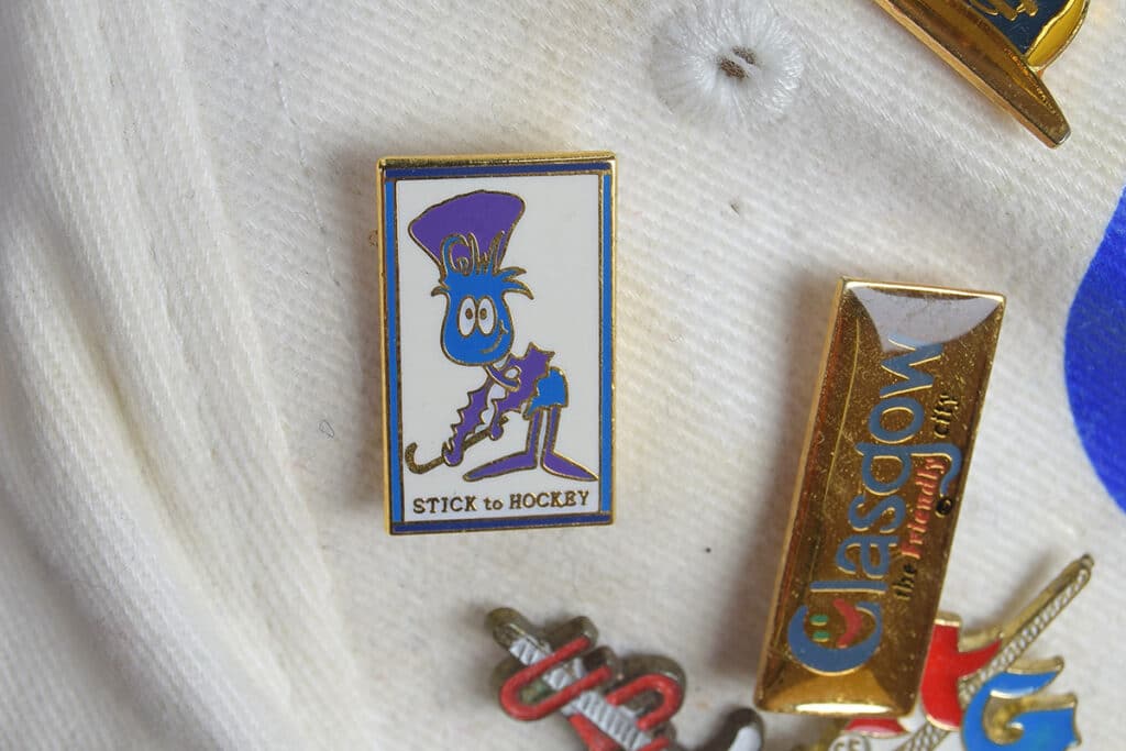 A small pin badge with a purple and blue cartoon character holding a hockey stick with the words "STICK TO HOCKEY" underneath