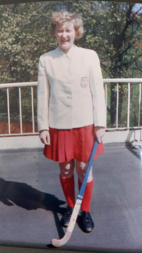 A picture of a woman with blonde curly hair in England hockey kit of white blazer with rose badge, white blouse, and scarlet red, knee-length skirt and socks.