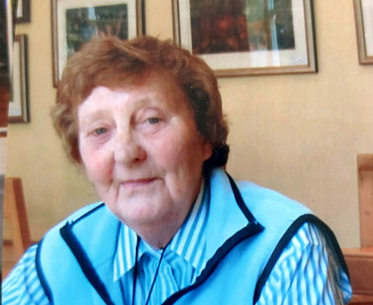 Photograph of Freda Walker. An elderly lady with reddish-brown curly hair wearing a light blue and white striped shirt and light blue sleeveless jacket.