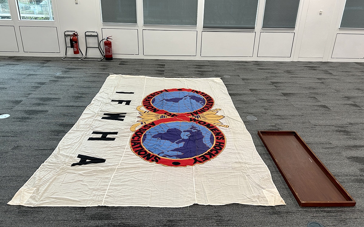 A large flag bearing the logo of the International Federation of Women's Hockey Associations is show fully laid out on the floor of an office. Its original display case, which is only about one-fifteenth on the size, is lying along side.