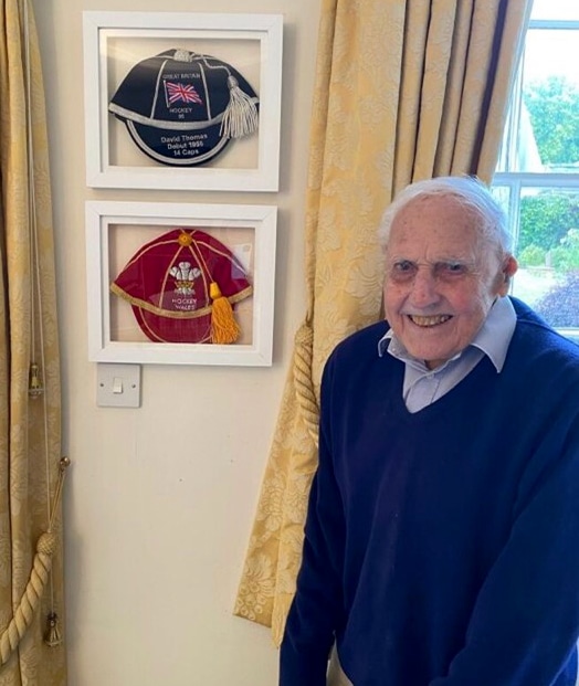 An elderly pale-skinned man in a royal blue jumper and light blue shirt stands to the right of centre smiling at the viewer. On the wall behind him, positioned between two sets of yellow curtains are two framed felt caps, one is navy blue with silver trim and tassel with the Union Flag logo, and the other is red with gold trim and tassel with the Welsh Prince of Wales logo of three white feathers behind a golden crown.