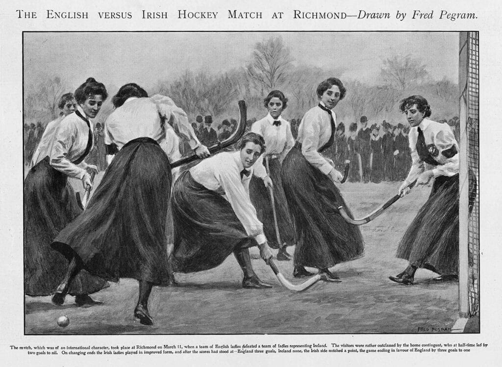 A black and white engraving in which seven women in various actions in competing for the ball during a hockey match. They have long dark skirts almost down to their angles fastened with a belt, dark stockings and black hockey shoes. Their torsos are covered by white blouses down their wrists and a black tie each. They have cloth badges with the emblems of their hockey teams sewn on to their breast pockets. Each wields a wooden hockey stick with a long hooked head. In the background a crowd of Victorian-era spectators, men and women, with hats, bonnets and mufflers form a line along the edge of the pitch.