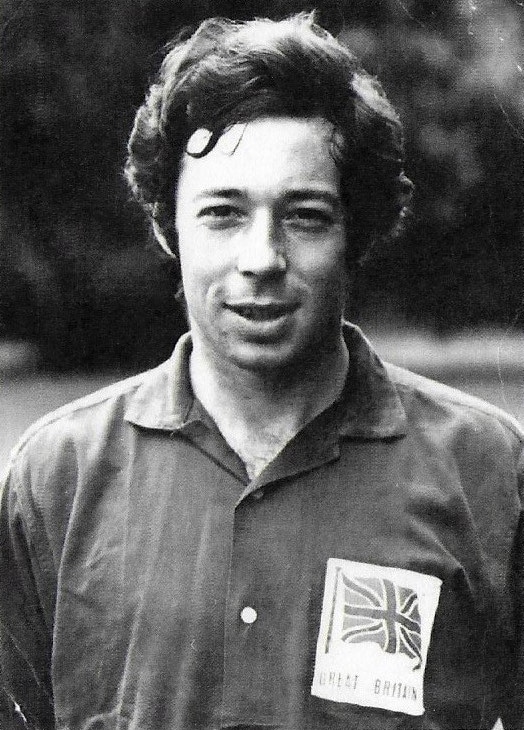 A black and white headshot photograph (face and upper torso) of a pale-skinned man facing the camera. He has dark, wavy hair which wraps around his ears joining with bushy sideburns. His lips are slightly parted as he smiles at the camera. He is wearing a Great Britain hockey playing shirt, grey in the photograph but red in reality; a cloth badge over his left breast pocket, white with a Union Flag design.