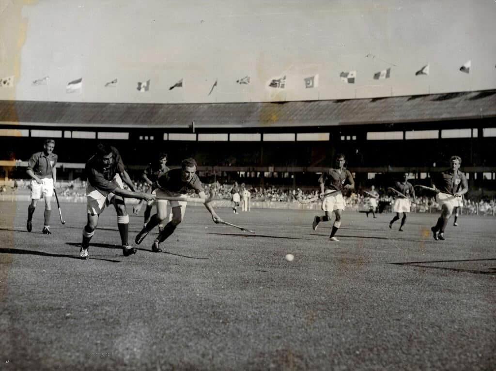 A black and white action photo of a men's hockey game in a large stadium, Great Britain vs Pakistan at the 1956 Olympic Games.