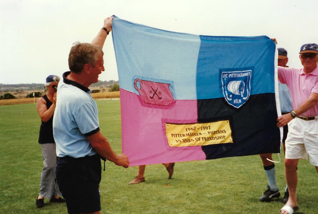 A colour image of two men holding a joint flag with the Puritan's colours (pink and blue ) and the Die Pittermänner Club colours (black and blue)