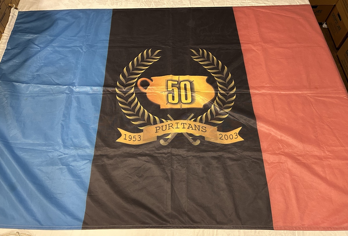 An image of a flag, with blue, black and red vertical stripes and a logo in the middle stating "1953 - 2003, Puritans"