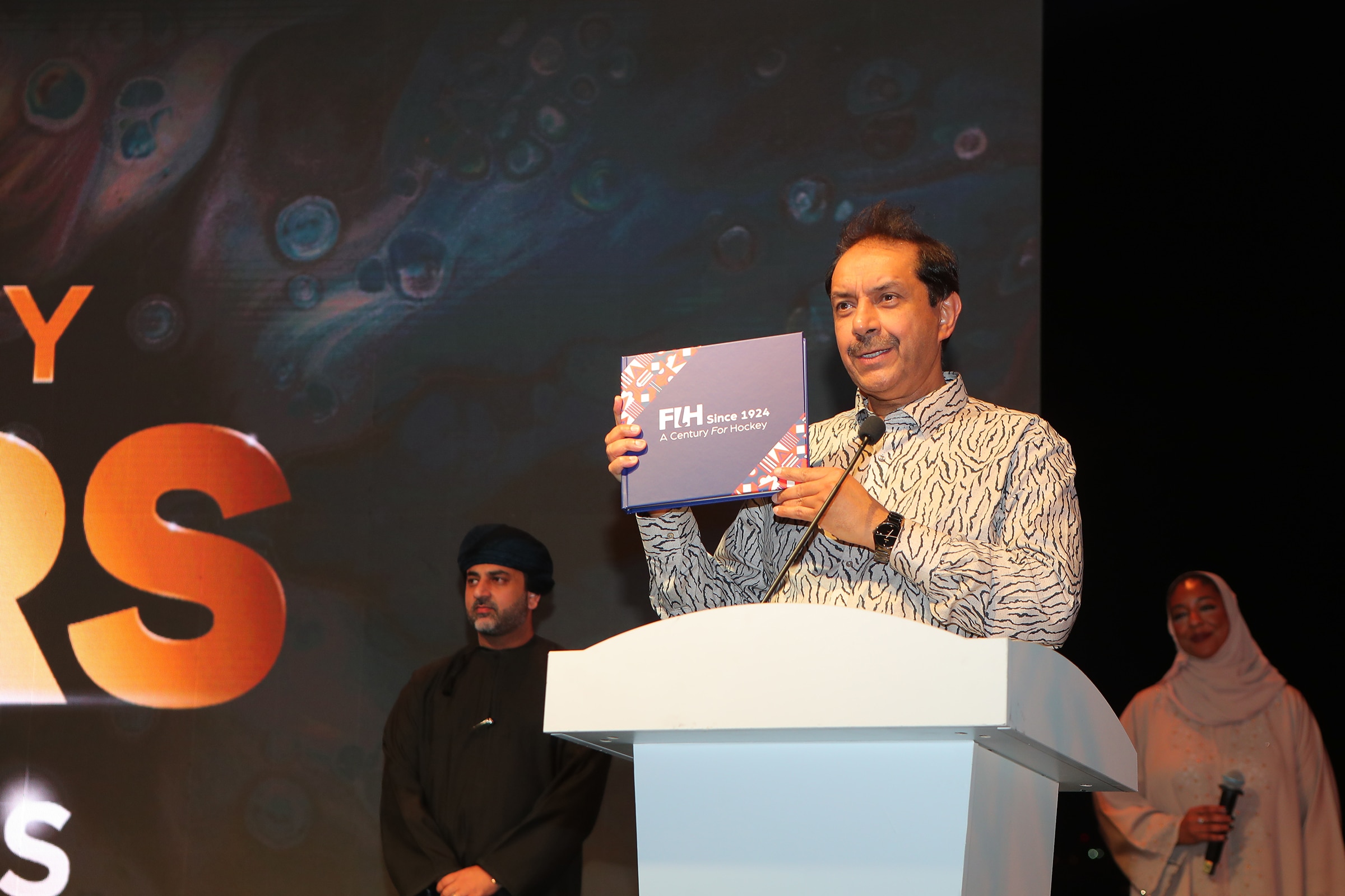 A man of South Asian heritage with short dark hair and a greying moustache stands behind a white lectern. He is wearing a white, long-sleeved shirt with black animal pattern and a black wrist watch. He is holding a copy of a large blue, red and white book. A man and a woman of Middle Eastern heritage look on in the background.