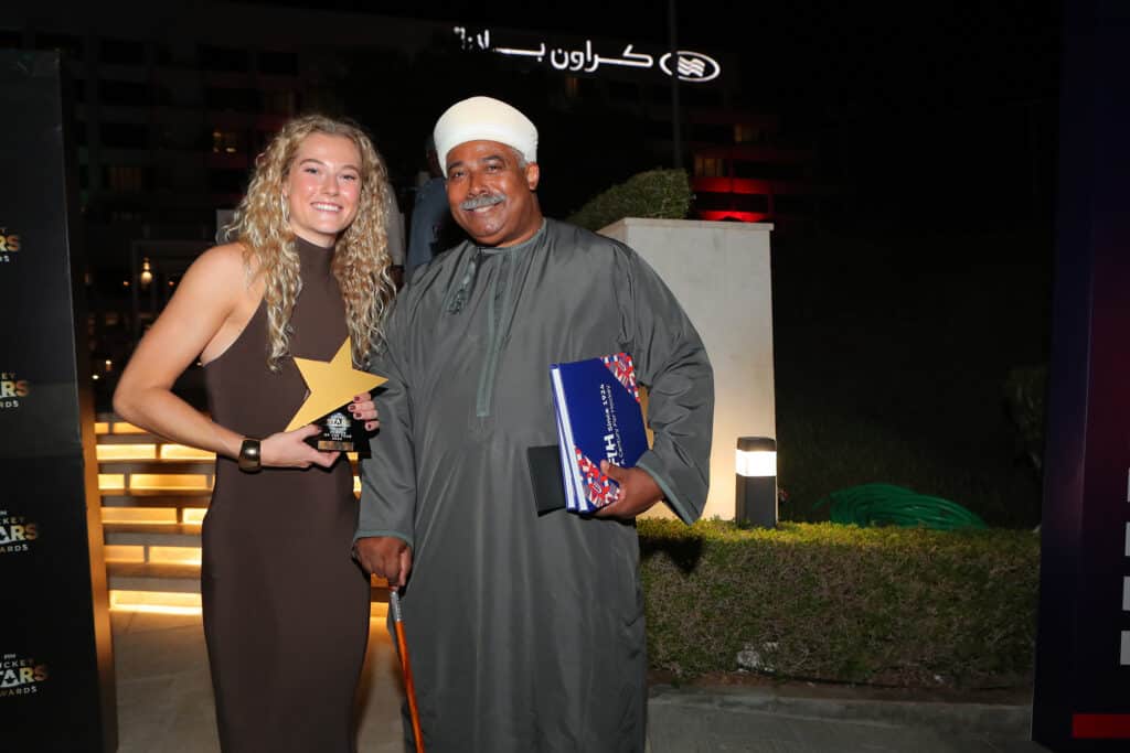 A young, Caucasian woman with long blonde hair wearing a long brown, sleeveless dress stands with an older Black man with a white moustache wearing a grey kaftan (a long-sleeved, ankle-length Islamic robe) and a white hat. She is holding a star-shaped trophy and he holds a walking stick cane in one hand and copies of a blue and red book under his other arm.