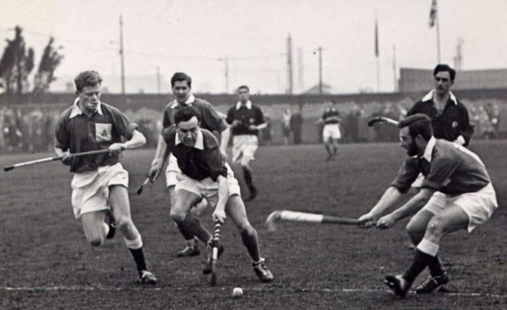 A black and white action photograph of a men's hockey match on grass. All players are wearing dark shirts with sleeves rolled up to the elbows, white shorts and boots and dark socks. A man on the right side of the photograph, bearded and wearing glasses, is attempting to tackle a player dribbling with the ball towards camera. A row of spectators is visible in the distance.