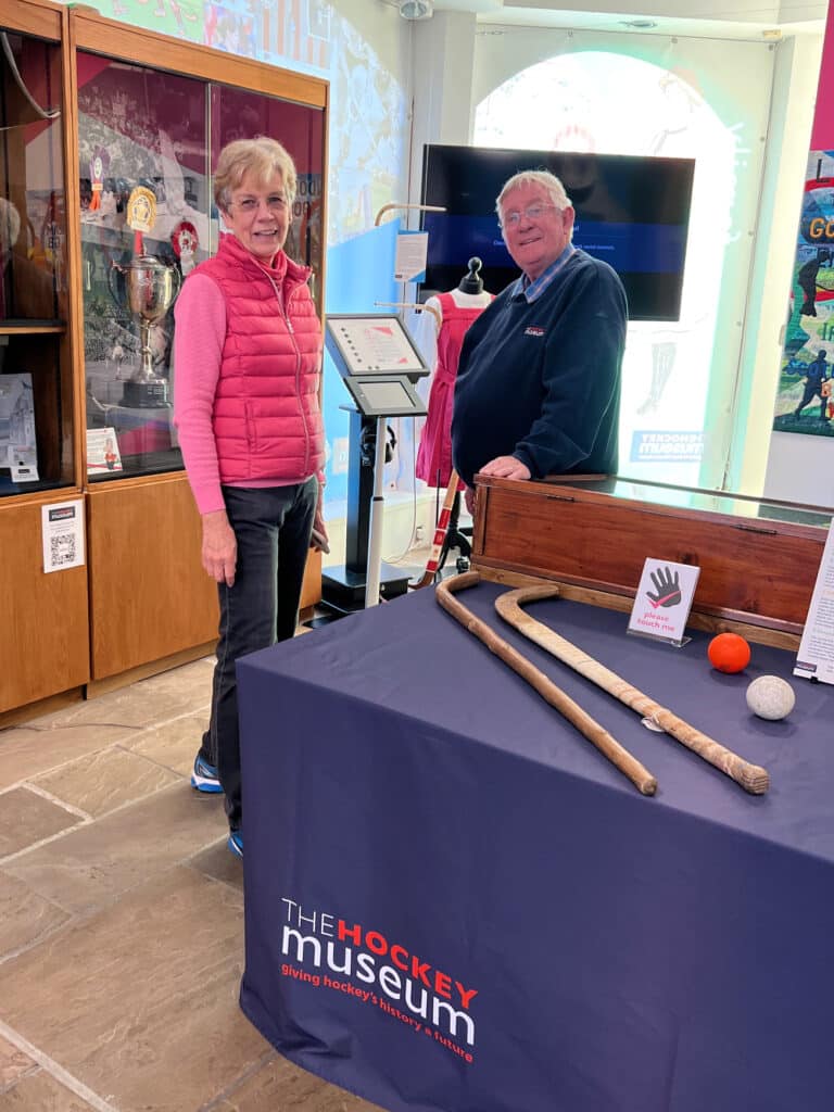 Two elderly people, a lady in a pink gillet and long-sleeved pink sweater with black jeans, and a man in a long-sleeved navy blue jumper and blue chequered shirt stand behind a table in a museum exhibition. Display cabinets and a screen are in front of large windows behind them.