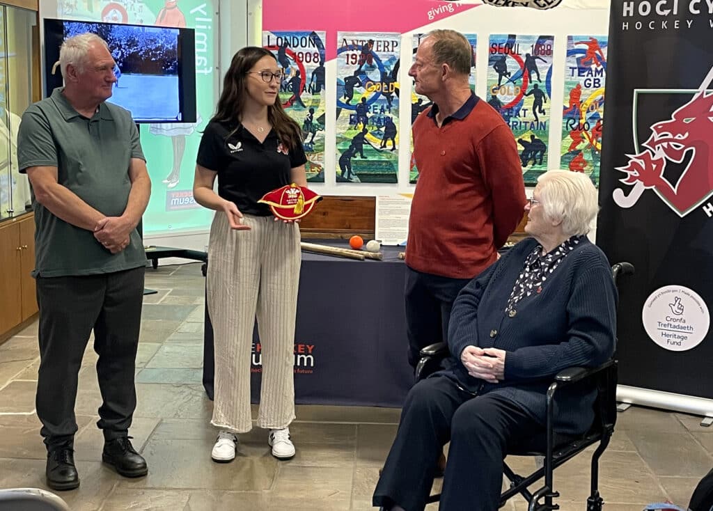 A man in a rust orange, long-sleeved jumper and jeans stands next to an elderly lady in a wheelchair. She in wearing a navy blue cardigan, white and navy blouse and navy trousers. Both are facing left towards two other people: a grey-haired man in a green polo shirt and blue trousers and a younger woman with long brown hair and glasses. She is wearing a black polo short and white, striped trousers and holding a red cap with yellow trim which she is about to present to the man in orange. A museum exhibition is the background.