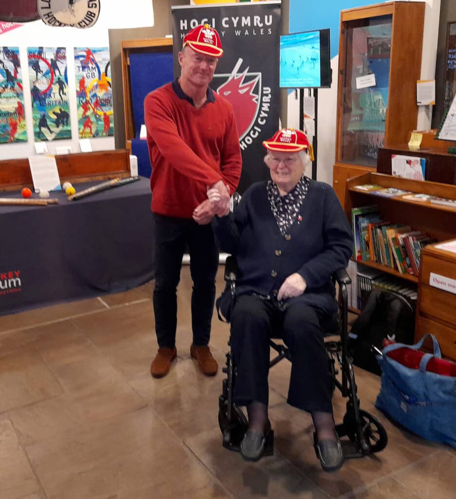 A standing man in a rust orange, long-sleeved jumper and jeans shakes hands with an elderly lady in a wheelchair. She in wearing a navy blue cardigan, white and navy blouse and navy trousers. Both are smiling at the camera wearing red caps with yellow trim.