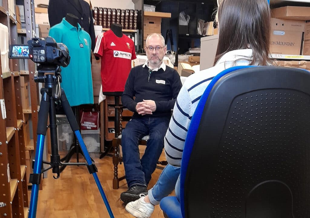 A grey-haired man in a navy blue, long-sleeved, collared shirt is being interviewed on camera. The camera and interviewer are in the foreground facing away from the viewer. Behind the man being interviewed are three Wales hockey shirts (red, turquoise and navy blue) on mannequins.