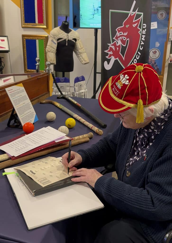 A elderly lady with white hair wearing glasses and a red cap with yellow trim is seated at a table covered by a navy cloth. She is signing various paper memorabilia. Also on the table are four hockey sticks and balls.