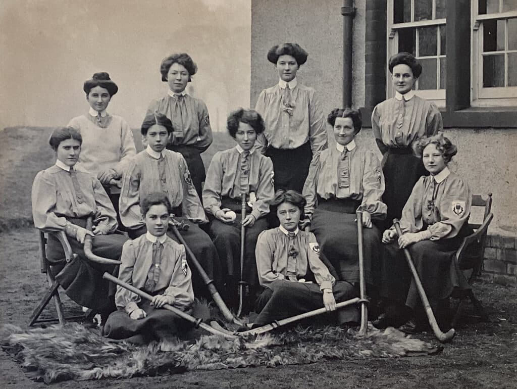 A black and white team photograph of three rows of Victorian-era women, the back row standing, the middle row seated on benches and chairs and the font row sitting on the floor. They hold old hockey sticks with long wooden heads. The all wear long-sleeved, loose-fitting blouses with high collars and ties, and long, dark skirts down to their ankles. Every woman's hair is tied or pinned up rather than hanging long. They are positioned in front of the corner of a house with a field behind.