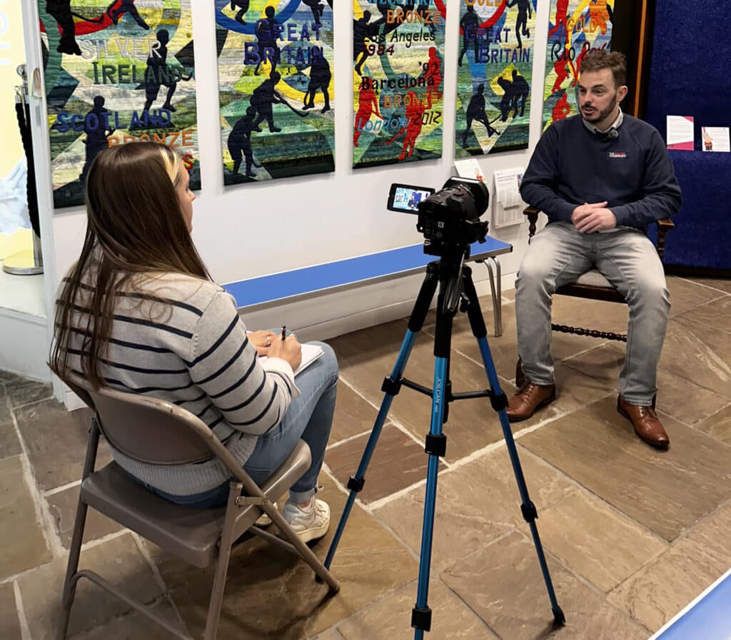 A young, bearded, brown-haired man in a navy blue, long-sleeved jumper and grey jeans is being interviewed on camera. The camera and interviewer are in the foreground facing away from the viewer. Behind the man being interviewed is a colourful, geometric quilt tapestry of Olympic hockey players.