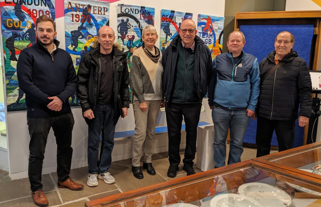 Five men and a woman gather in a room with a stone-tiled floor behind a display case and in front of a colourful series of tapestries, facing the camera.