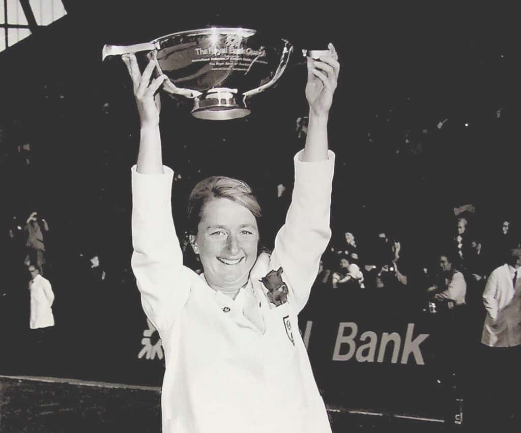 A black and white photograph of a woman in a white England blazer with pocket badge of roses lifts a bowl-shaped trophy above her head. A crowd cheers in the background.