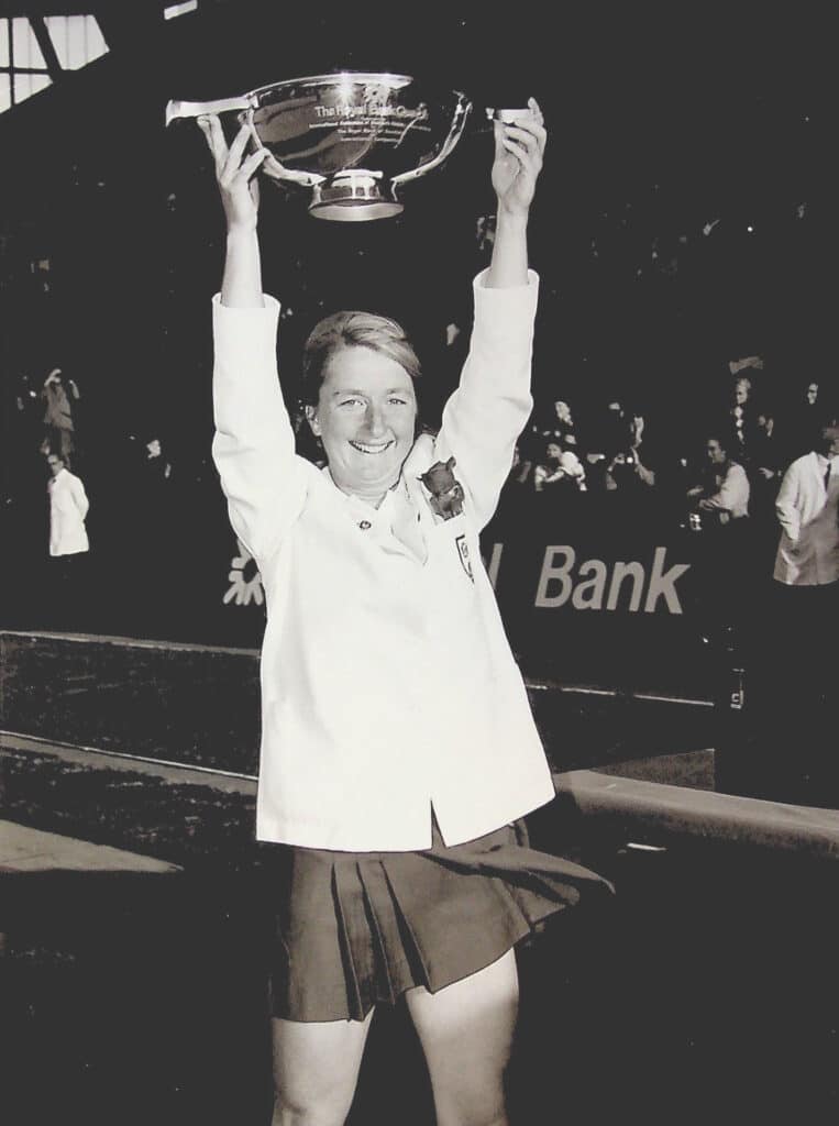 A black and white photograph of a Caucasian woman in a white England blazer with a roses badge and a dark skirt lifts a bowl-shaped trophy above her head. A crowd cheers in the background.