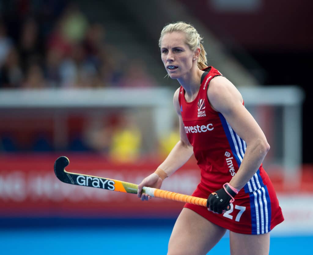 A colour image of a female hockey player. She has blonde hair tied up in a pony tail, and wears a red Great Britain sleeveless top and skirt with blue and white side stripes. Her hockey stick is black and orange with an orange grip.