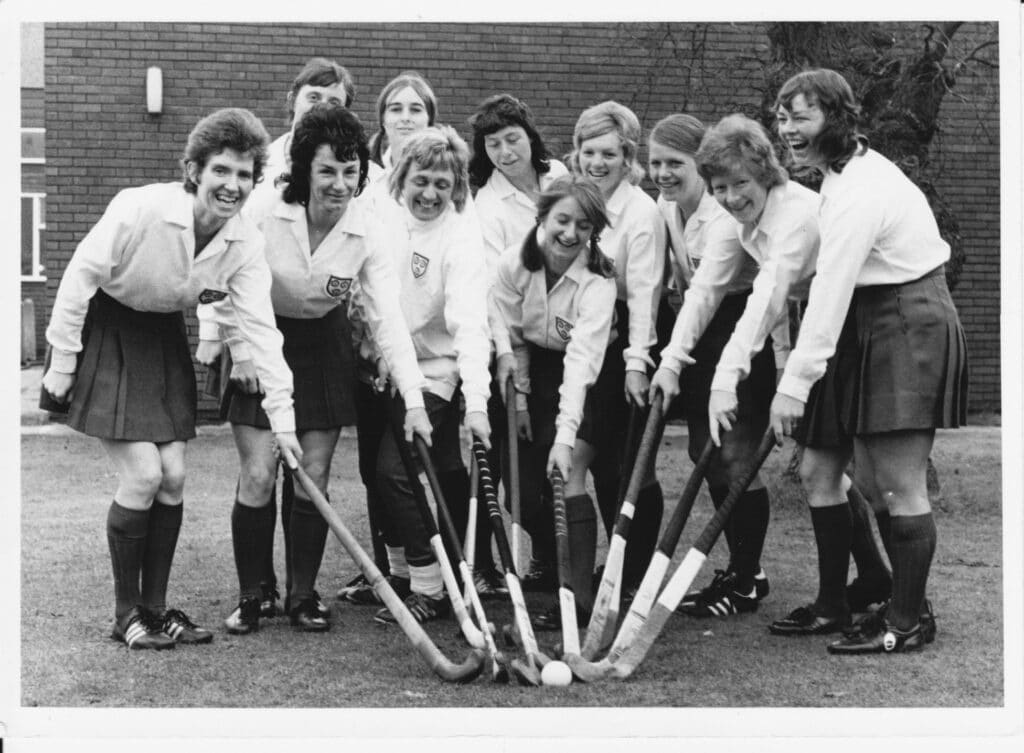 A black and white photograph of the 1974 England women's team. They players are all leaning into a huddle joining their hockey sticks at the centre point of the photograph. They are wearing white shirts or jumpers and dark skirts and socks.