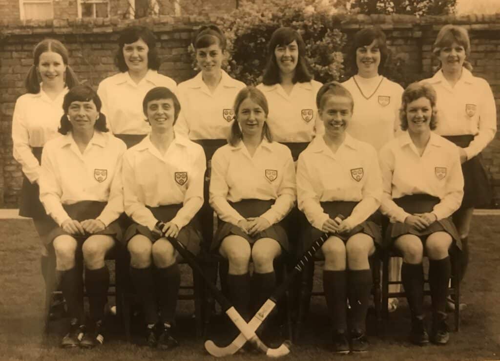 A sepia-coloured, black and white image of an England women's hockey team. The players are in two rows, the back row standing and the front row seated. The players wear white shirts and dark skirts and socks.