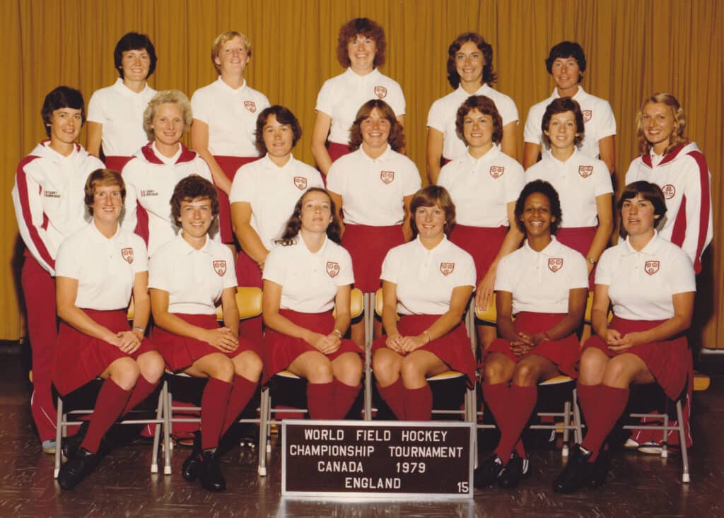 A colour image of the England women's hockey squad, manager and physiotherapist looking at the camera. They are in three rows: the back two rows standing and the front row seated. The players are wearing white polo shirts and cardinal red skirts and socks. The two staff are in white tracksuit tops with red sleeve stripes and red tracksuit bottoms.