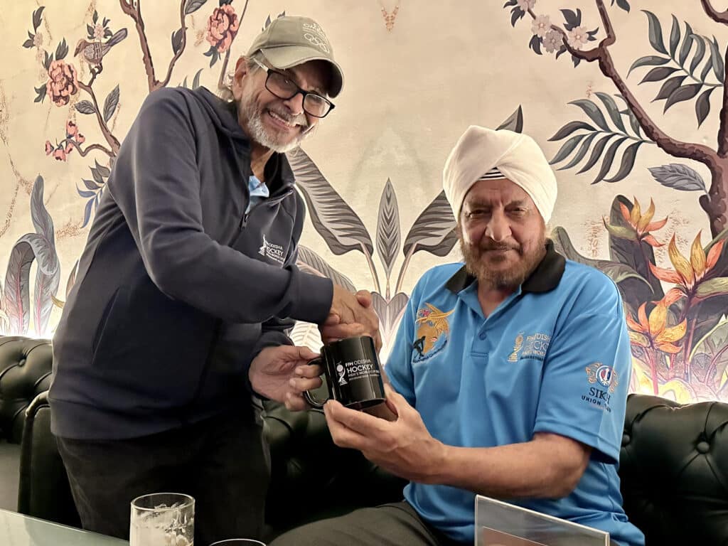 An image of two men of South Asian heritage shaking hands looking at the camera. The left man is standing. He is has a short, grey beard and glasses and is wearing in a blue jumper and khaki cap. The man on the right is seated. He has a light brown beard and is wearing a cream-coloured turban and a light blue polo shirt.