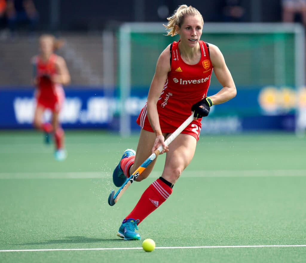 A colour photograph of a female hockey player in action, dribbling with the ball towards camera. She has blonde hair tied up in a pony tail, and wears a red England sleeveless top, skirt and socks. Her hockey shoes and light blue and her stick light blue and orange with a white grip.