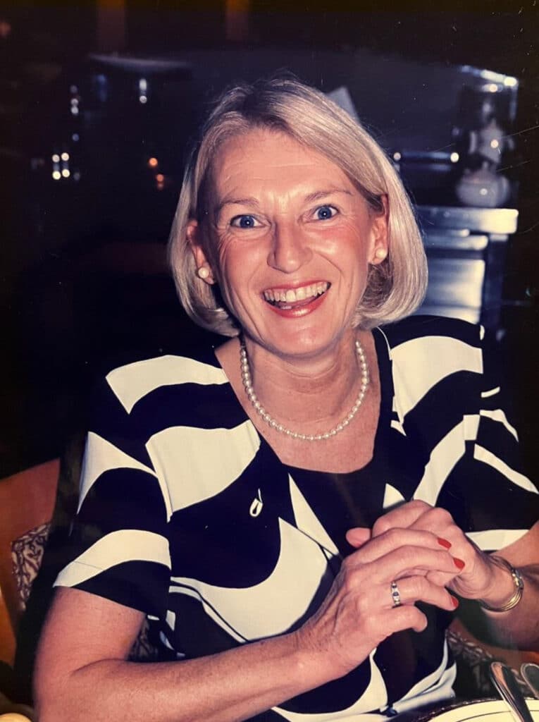 A colour picture of a blonde-haired lady smiling at the camera. She is wearing a black and white blouse with a pearl necklace.