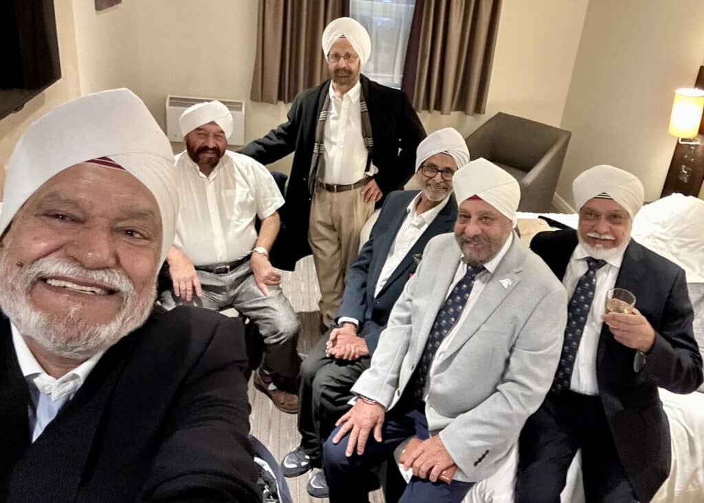 A group of six South Asian heritage men are seated or standing in a hotel room. They are smartly dressed in suits and white turbans, taking a selfie.