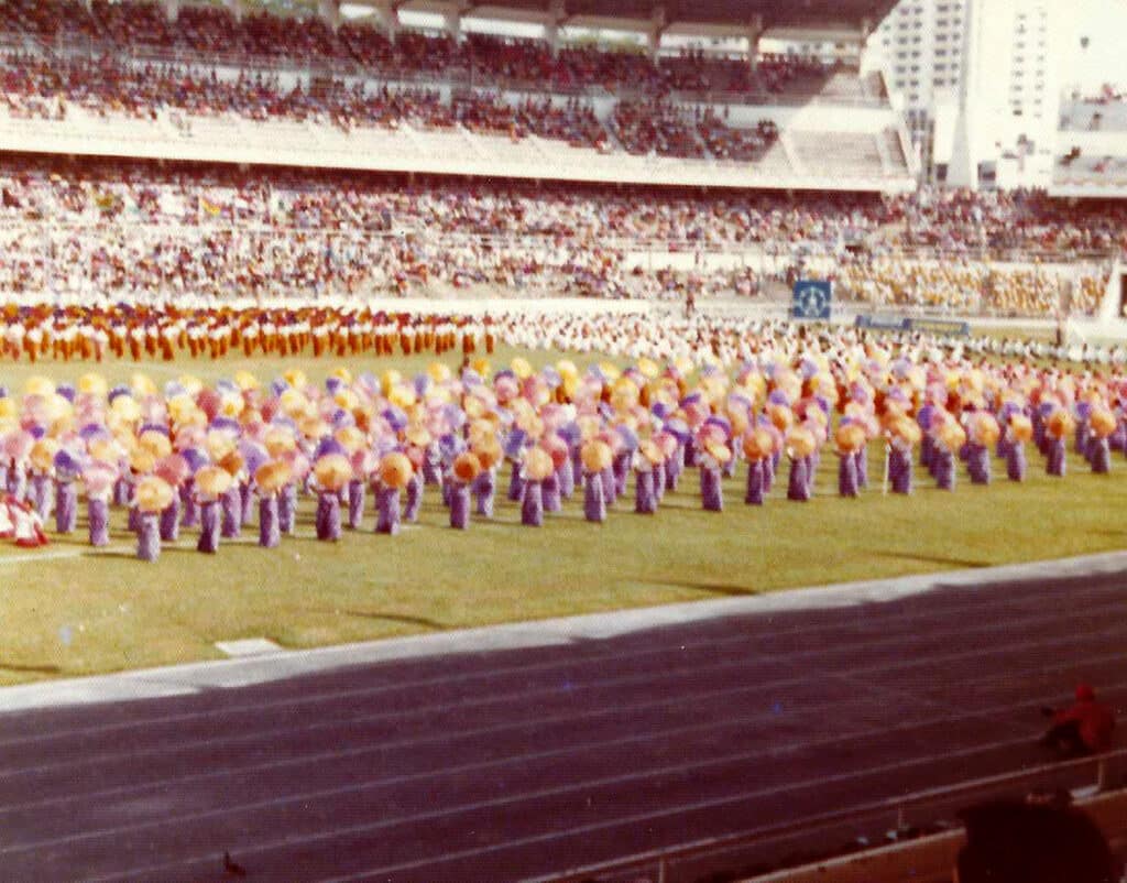 Landscape photograph of the inside of a stadium, crowds of people are in the stands with performers from a celebratory ceremony in the middle of the grass pitch, which is surrounded in the foreground by the stadium athletics track. The scene is a riot of colour.
