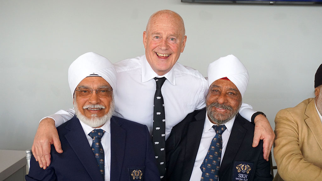 Two elderly Sikh men of Indian heritage, both seated, pose with an elderly, blad Caucasian man who is standing between them with his arms around their shoulders. The Caucasian man is wearing a white shirt and navy blue tie with white pigs on it. The Sikh men are both in dark jackets with Sikh Union Club pocket badges and blue ties. They are wearing white turbans.