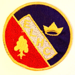 A circular cloth badge with yellow border dissected diagonally in half from top left to bottom right. The lower left half is red with a yellow tree and the top right half is navy blue with a yellow crown. The initials E L H C divide the middle, yellow letters on a white background.