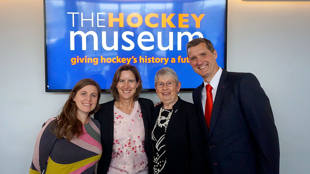 Four people, all Caucasian, three women and a man, pose in front a large TV screen with The Hockey Museum logo on it. The left-most woman has long brown hair and is wearing a multi-coloured patterned dress. The left-centre woman has shoulder-length brown hair and is wearing a black jacket over a light pink flowery top. The right-centre woman is has short grey hair and glasses. She is wearing a black jacket over a black and white dress with a chunky white necklace. The man is very tall with short brown hair. He is wearing a navy blue suit with white shirt and a red tie.