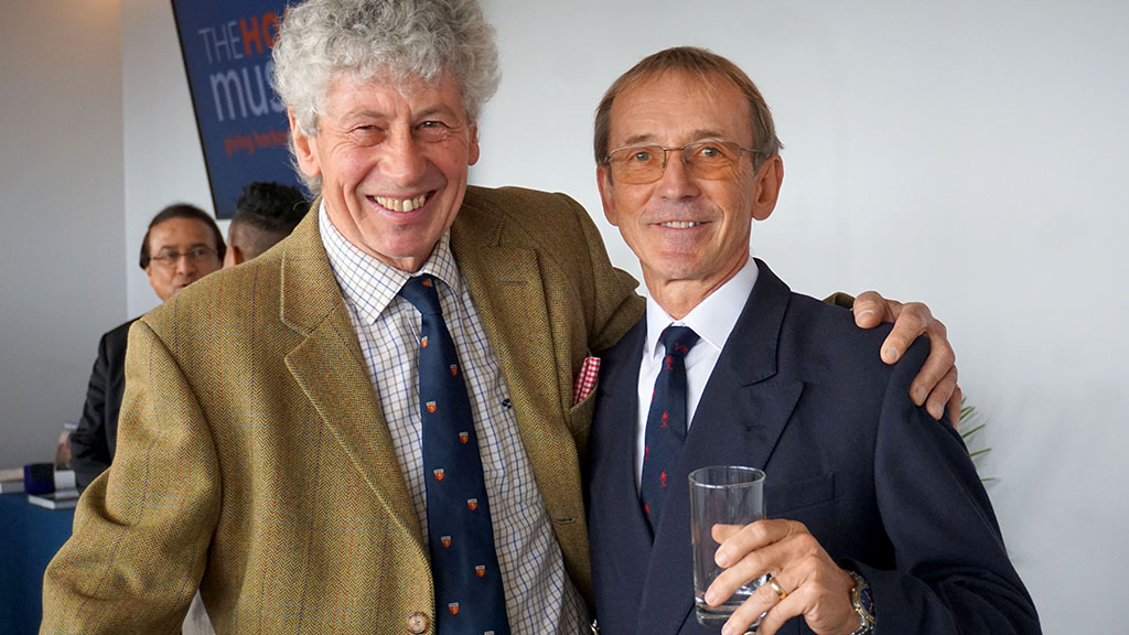 Two Caucasian men wearing suits face the camera smiling, arms clasped around each other's shoulders.