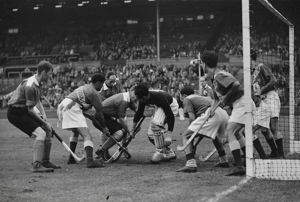 Hockey players crowd around the goalkeeper in the goal mouth during a hockey match. Pakistan's players are defending against Great Britain. There is a large crowd in the background.