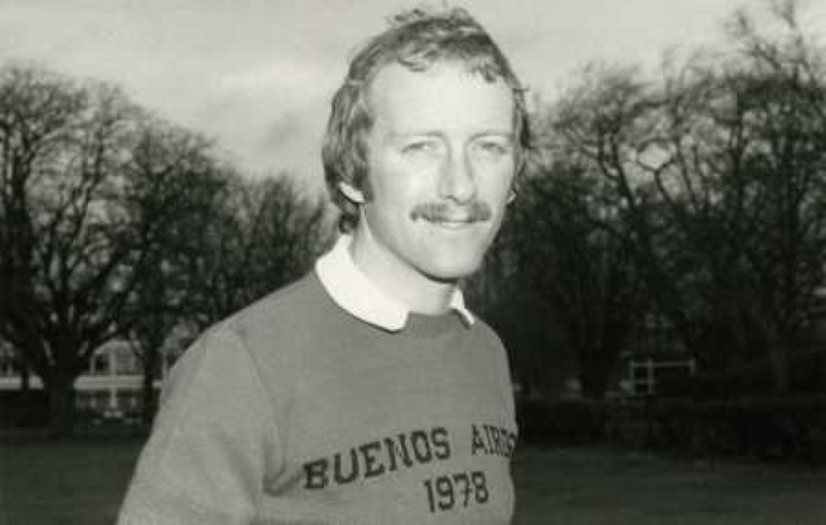 A black and white image of a young man with a moustache in a sweater looking at the camera