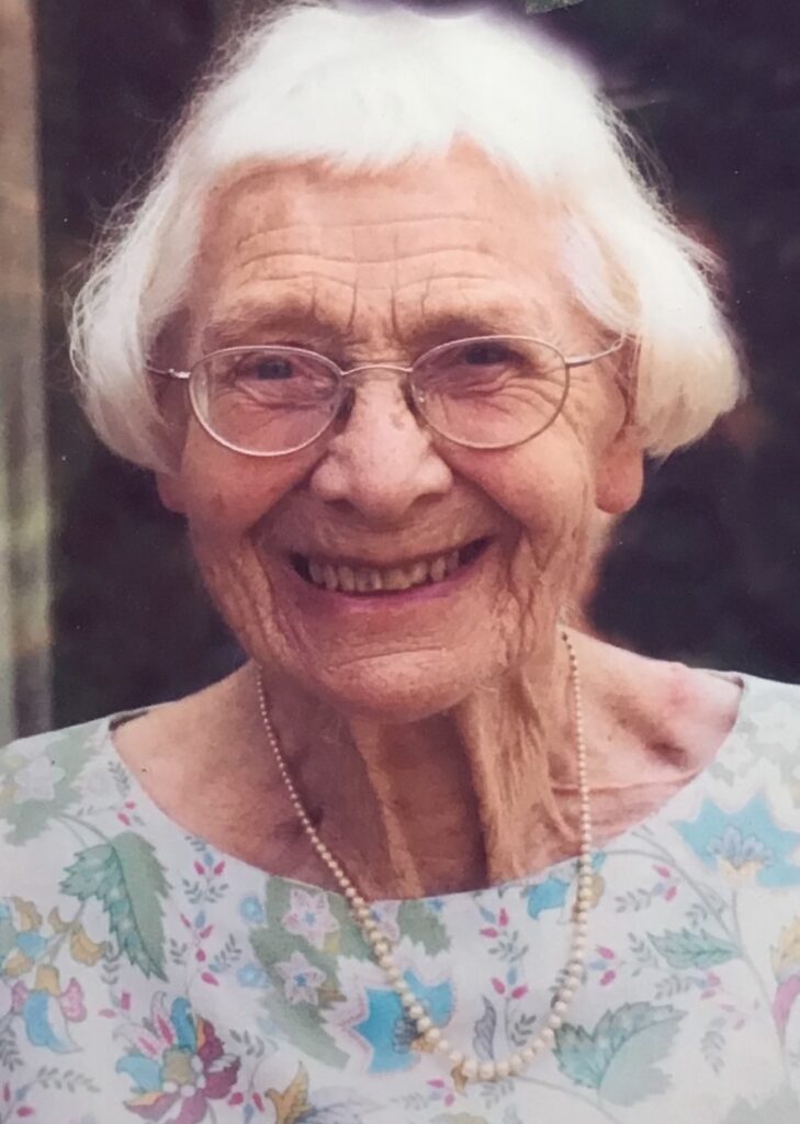 A colour picture of an elderly lade in a floral dress smiling at the camera