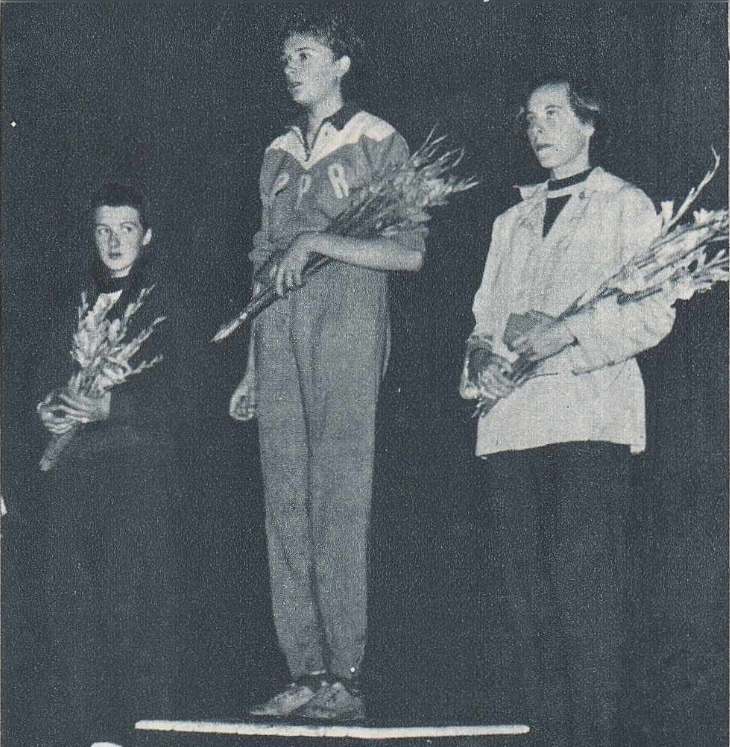 A black and white photo of three women in ahtletic tracksuits on podiums holding flowers