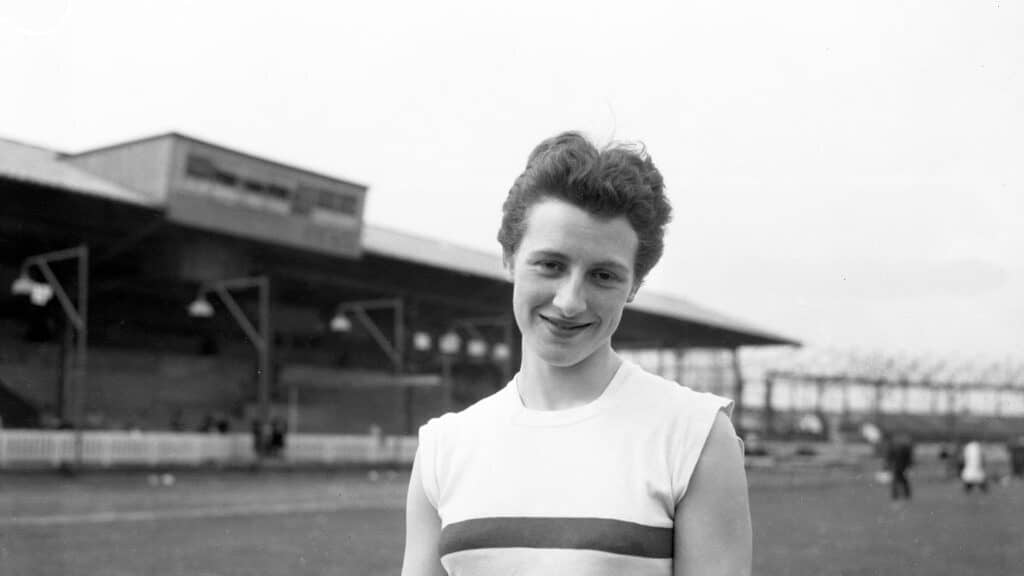 A black and white photo of woman in atheltics kit looking at the camera in a stadium