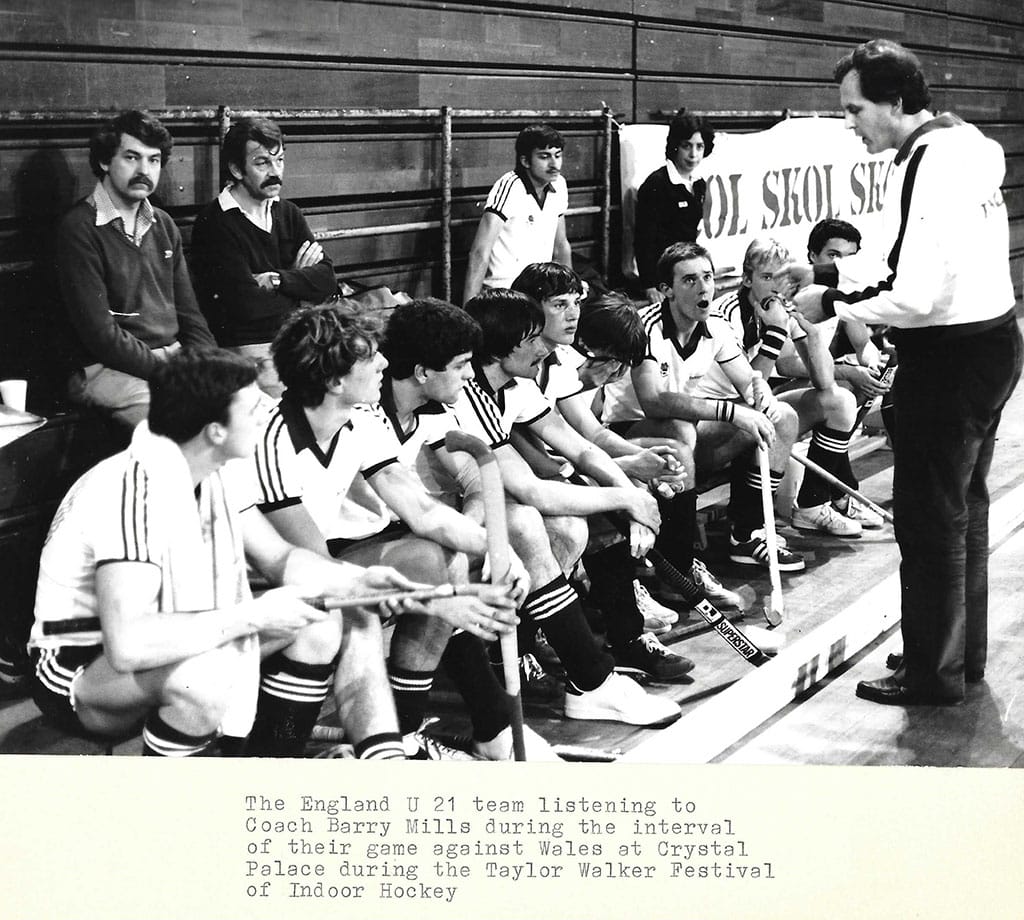Black and white photograph of a group of young men in white tops and dark shorts and socks. They sit in a row, diagonally from bottom left of the photograph to top right, looking up to a standing man who is giving a team talk.