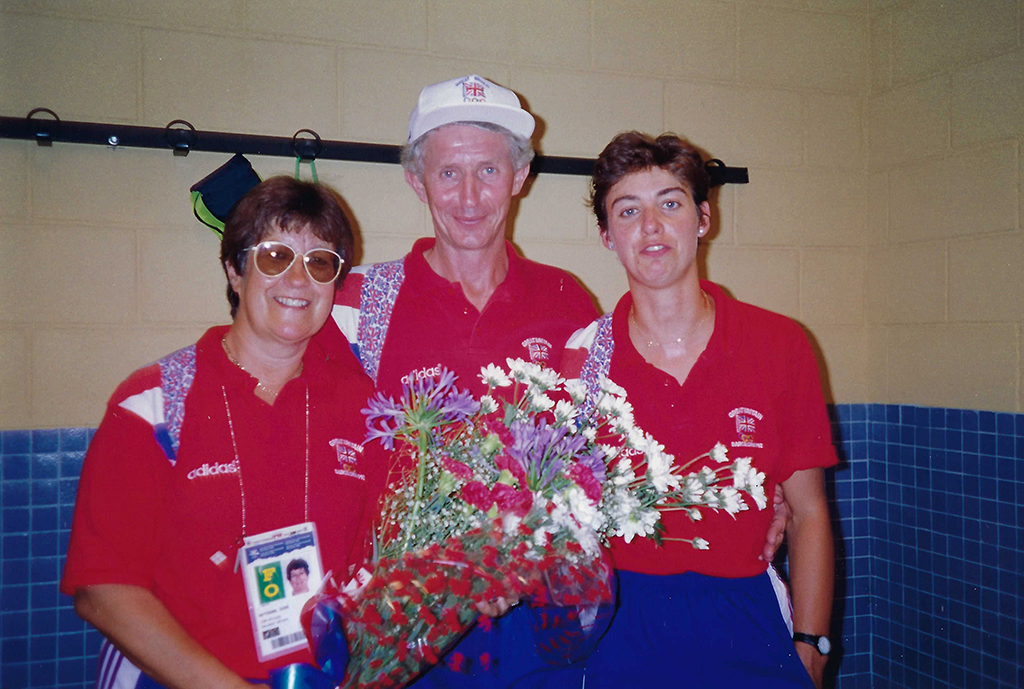 A tall middle-aged man with grey hair wearing a white cap with the Union Flag stands between two women in a blue-tiled sports changing room. The left woman is also middle-aged with short brown hair and sunglasses; the other woman is younger with short brown hair. They all wear the same Adidas uniform: red polo shirt with a Union Flag stripe on the right side, and royal blue shorts with white stripes.
