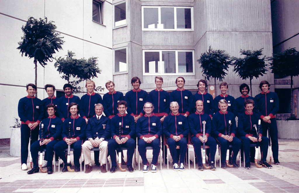 Team photograph of two rows of men, the front row seated. They are wearing navy blue tracksuits with red collars, cuffs and zip. The men are positioned in the courtyard of an austere concrete building, between them and the building are five small trees in large concrete pots.