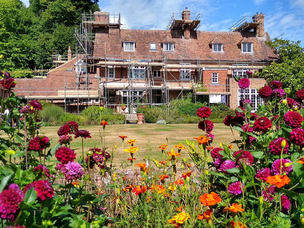 A red brick and red tiled house is surrounded by scaffolding. In the foreground flowers of purple, red, pink and orange frame the view to the house.