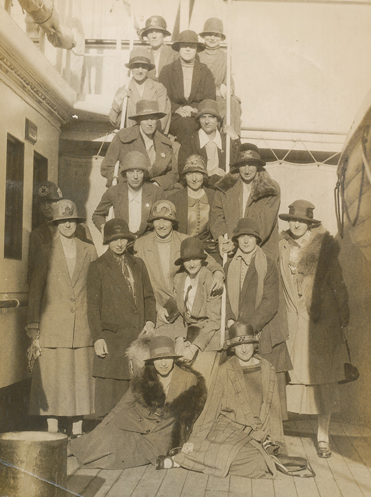 Sepia photograph of a group of women in coats, blouses, big coats and large sun hats on the deck of boat. Some stand posed on the stairs between decks; others lounge or stand at the foot of the steps.